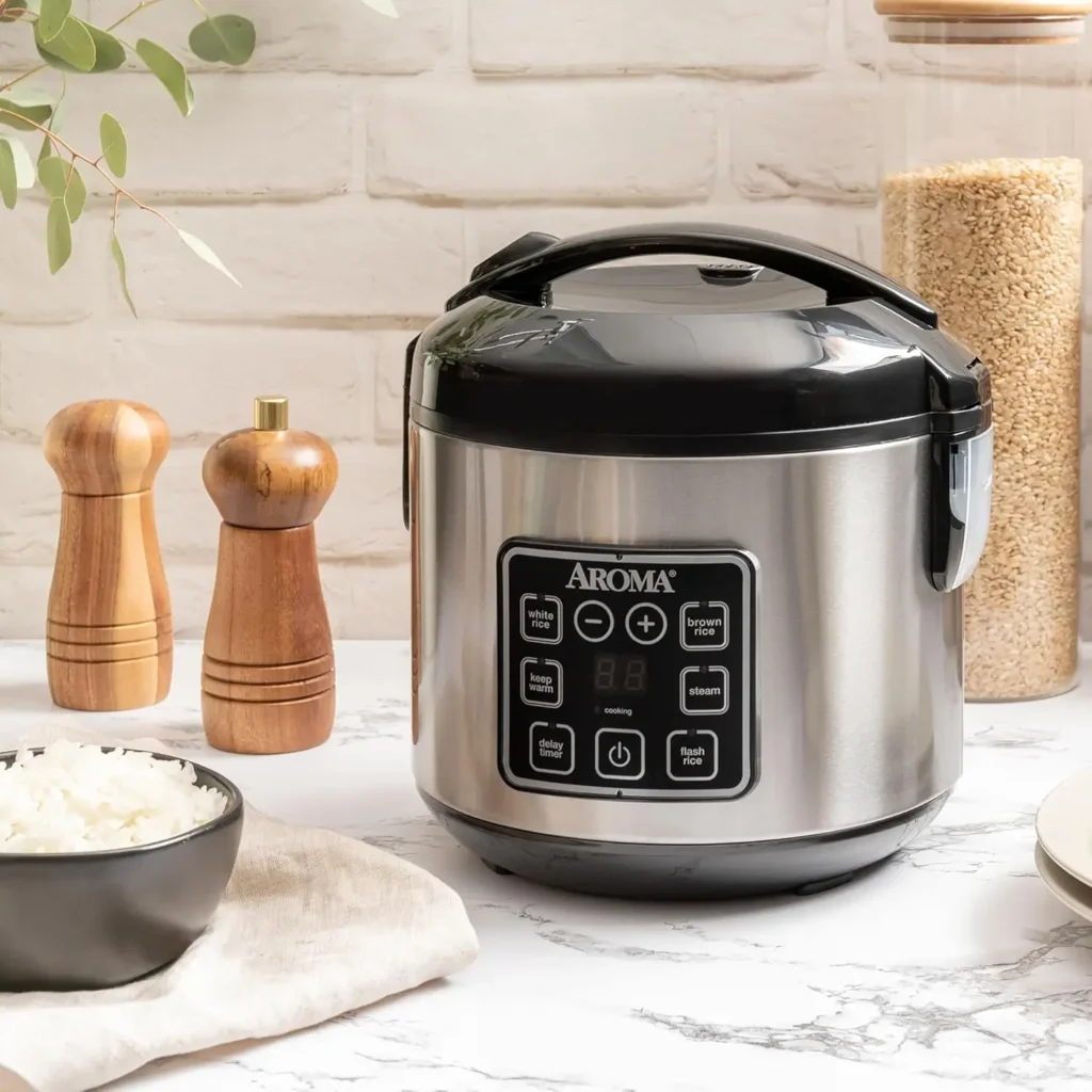 AROMA ARC-914SBD stainless steel rice cooker placed on a marble kitchen countertop next to a bowl of freshly cooked white rice, wooden salt and pepper mills, and a glass jar of uncooked rice
